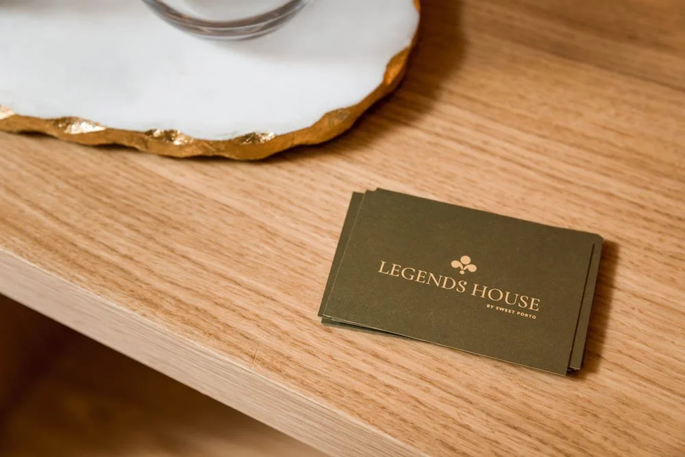 Cartões de visita do Legends House, com fundo verde escuro e texto dourado, sobre uma mesa de madeira.