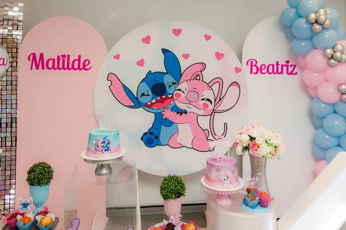 Decoração de festa infantil com tema de personagem do desenho 'Lilo & Stitch'. Fundo decorado com ilustração de Stitch e Angel, duas personagens fofas. Dois bolos decorados em tons de rosa e azul, com detalhes relacionados ao tema. Flores e balões em