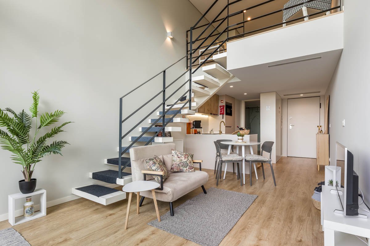 Sala de estar moderna com piso de madeira, sofá bege com almofadas decorativas, mesa de centro branca, planta em vaso preto, escada de madeira com corrimão de metal preto, mesa de jantar com quatro cadeiras, cozinha ao fundo, porta de entrada, televi