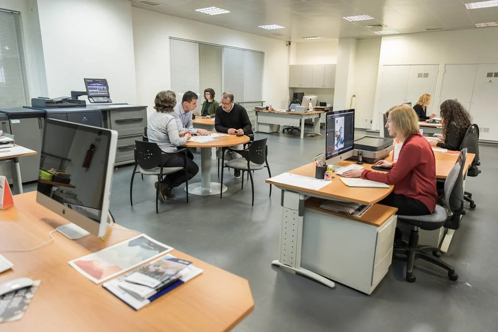 Pessoas trabalhando em um escritório com computadores, incluindo uma equipe em uma reunião ao redor de uma mesa e outras pessoas usando computadores individuais