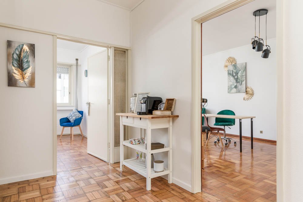 Vista de interiores de escritório com piso de parquete, paredes brancas, decoração com quadros e móveis distintos em cada sala, uma com uma cadeira azul e outra com uma mesa e cadeira verdes.
