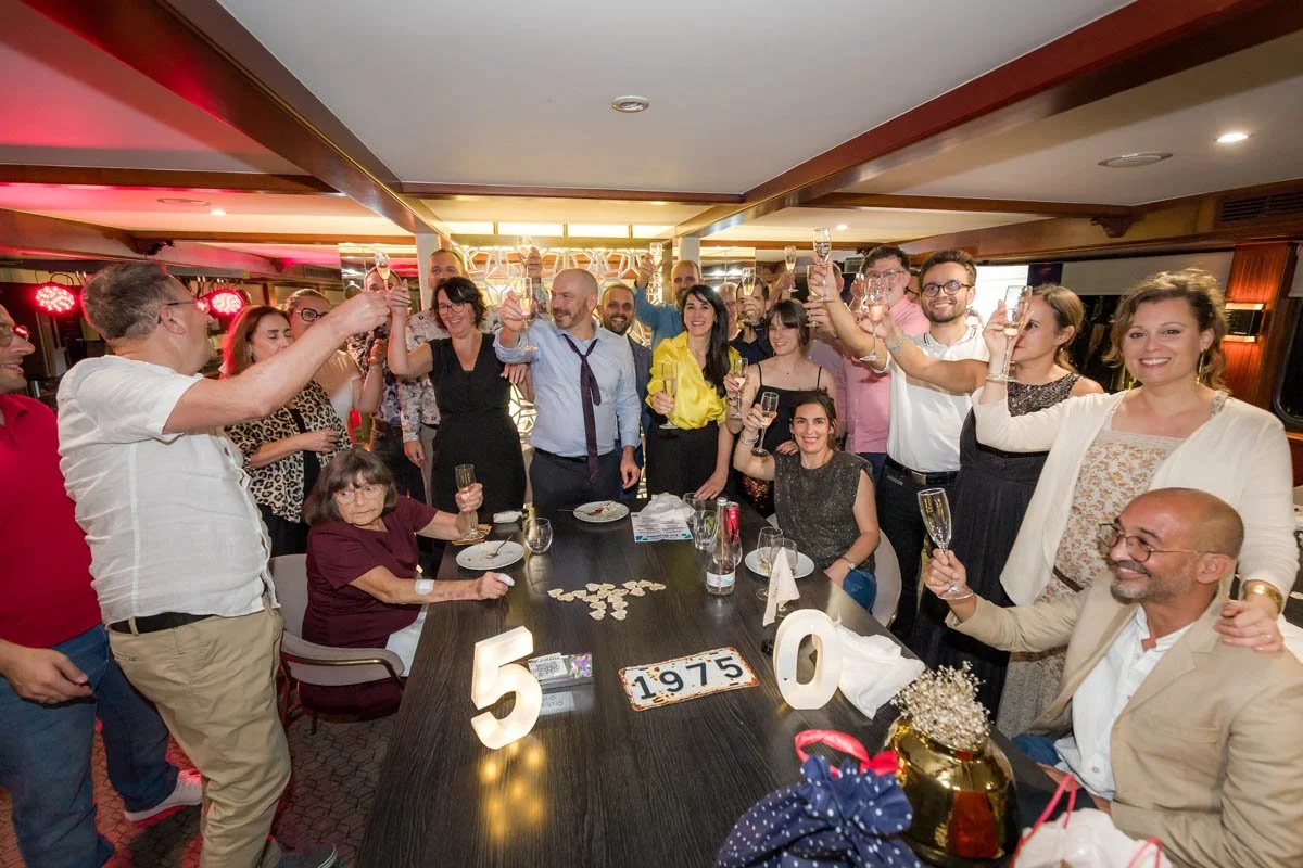 Grupo de pessoas celebrando ao redor de uma mesa, com taças levantadas em brinde, em uma comemoração de 50 anos de casamento, com decoração de 50, 1975 e elementos de festa na mesa.
