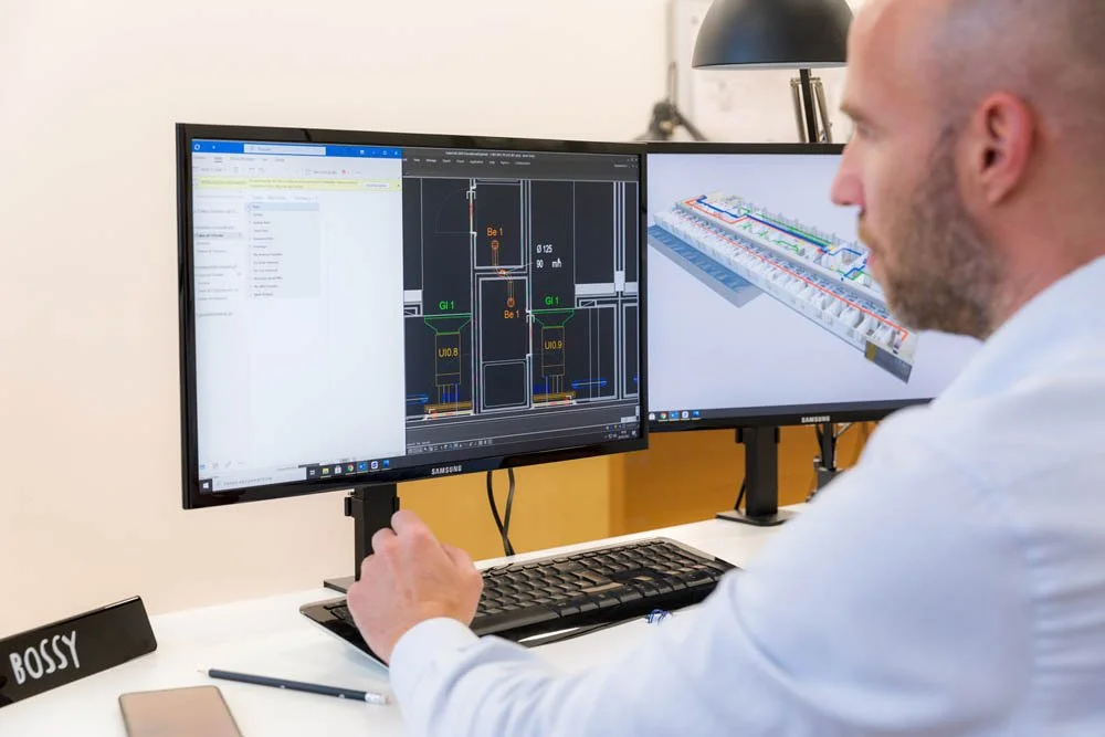 Homem trabalhando em computador com dois monitores, um mostrando um projeto técnico e o outro uma modelagem 3D, em um escritório.