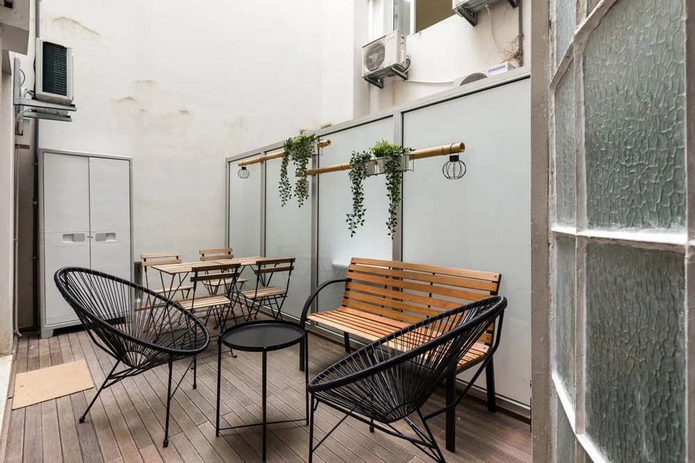 Varanda com poltrona de fibra, mesa pequena, banco de madeira, mesa de jardim com cadeiras, parede com plantas penduradas e aparelhos de ar-condicionado.