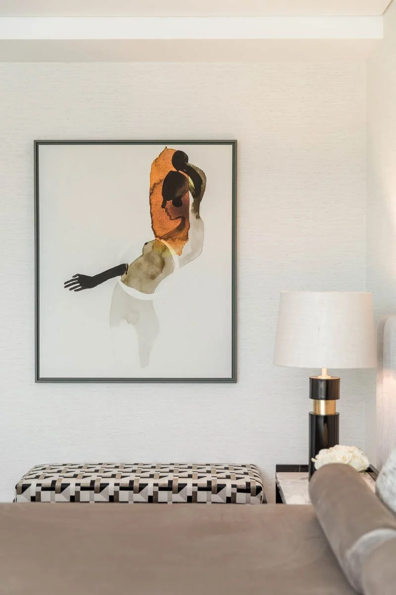 Quadro abstrato de uma mulher com cabelo laranja e pele escura, posicionada na parede de um ambiente decorado com uma mesa lateral com abajur e banco com estampa geométrica.