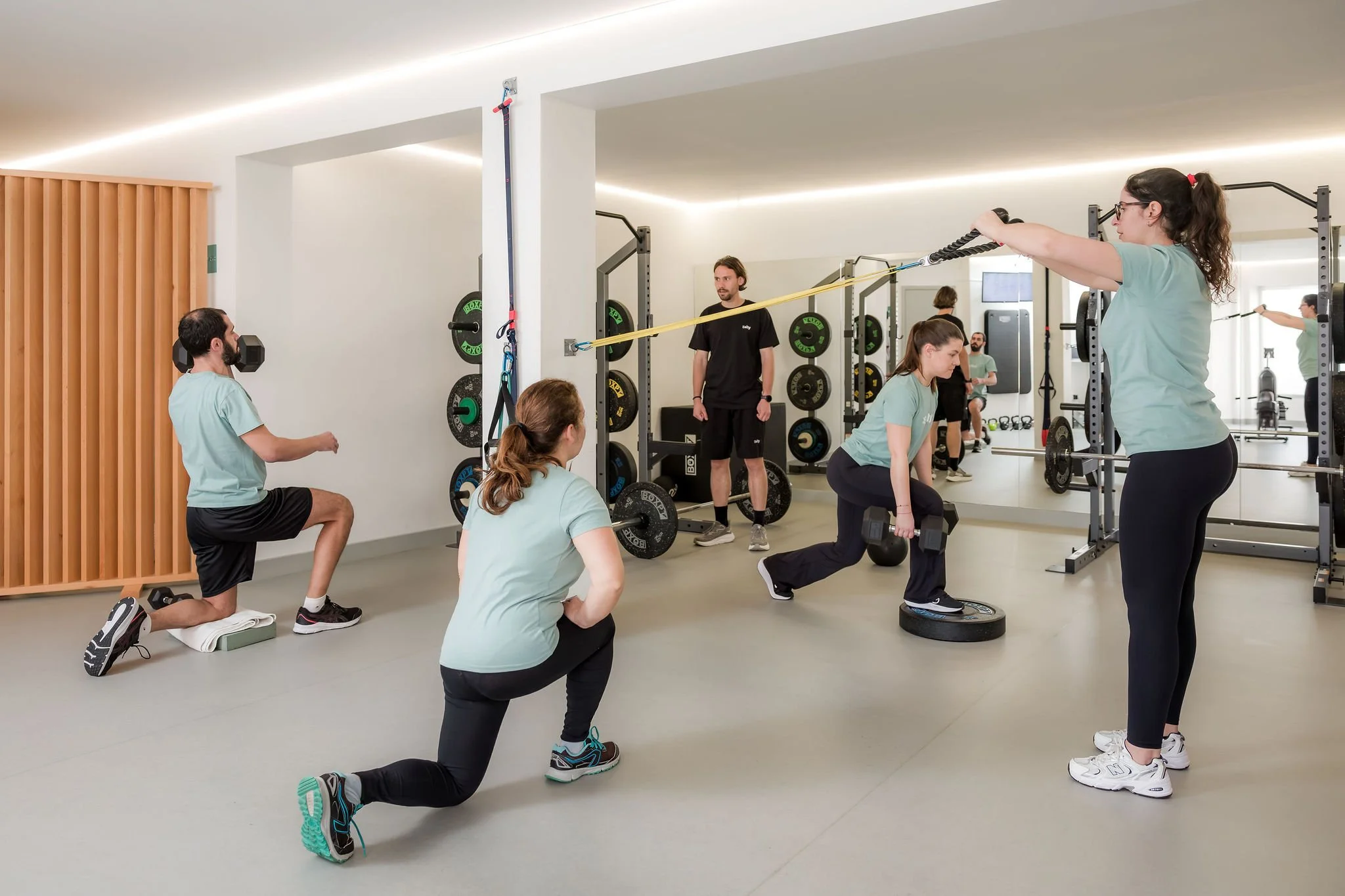 Pessoas fazendo exercício em uma academia, incluindo agachamentos, levantamento de peso e treino com faixas de resistência.