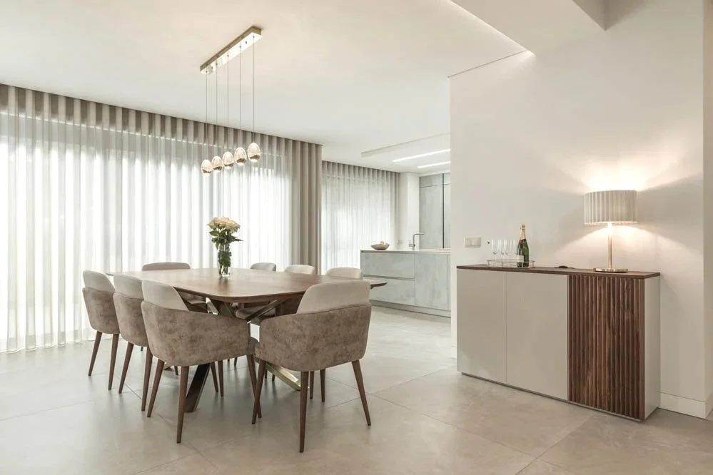 Sala de jantar com mesa de madeira e oito cadeiras de estofado claro, iluminada por luz natural de cortinas transparentes, com uma parede branca e uma área de cozinha ao fundo, incluindo armários brancos, uma pia e uma bancada, decorada com um aparador de madeira com lâmpada e garrafas na superfície.