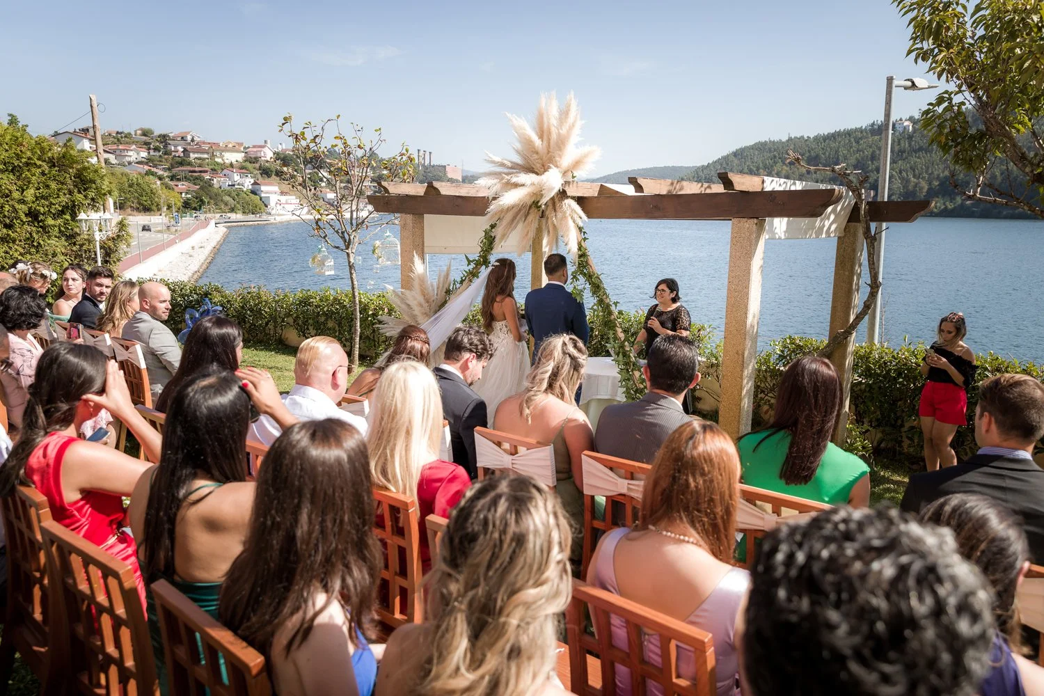 Casamento ao ar livre com vista para um rio ou lago, com uma cerimônia sob uma estrutura de madeira decorada com folhagens e pampas, na presença de convidados sentados em cadeiras de madeira, em um dia ensolarado.