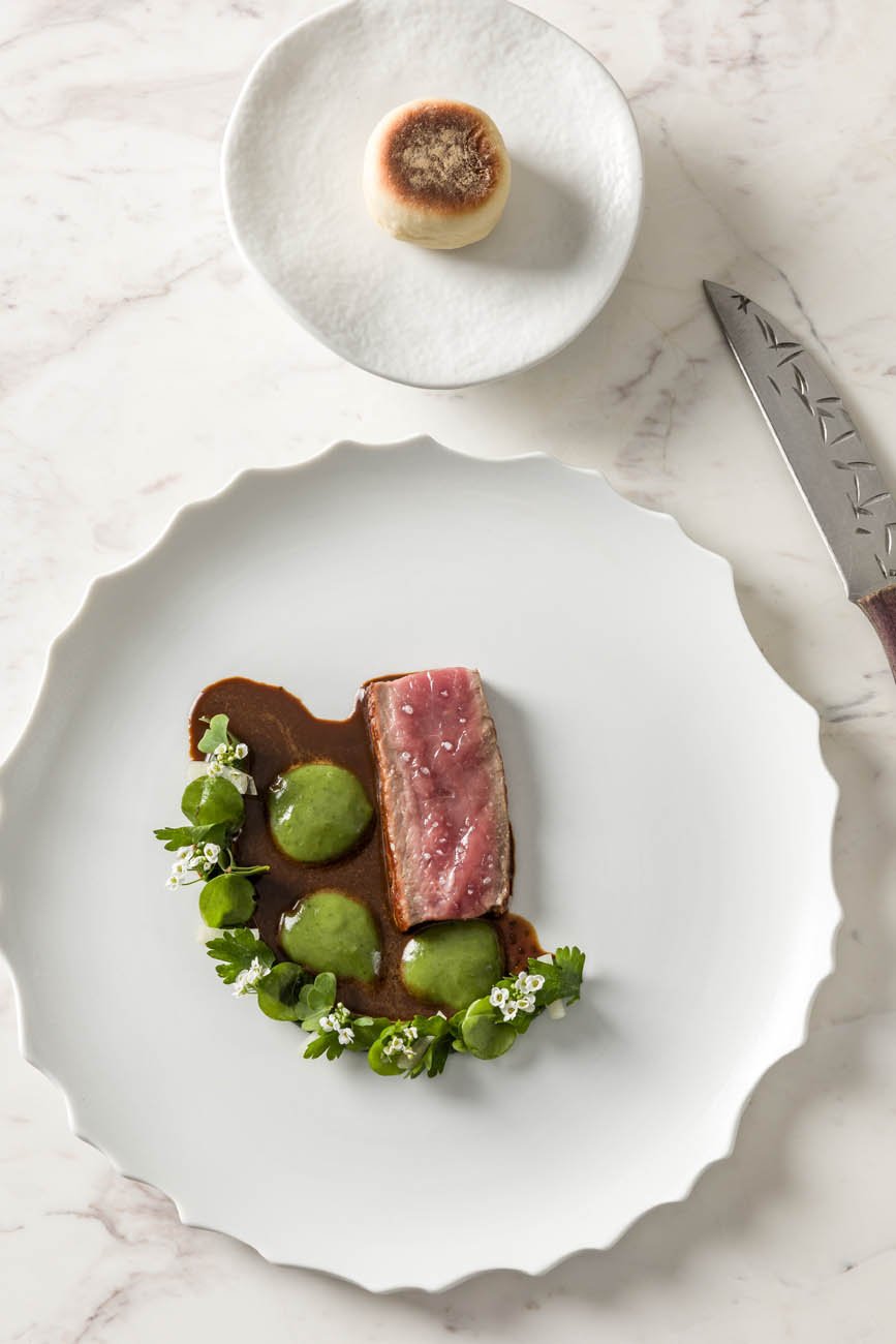 Prato de carne com molho, acompanhada por geleias verdes decoradas com ervas e flores, numa louça branca com borda ondulada; sobremesa de pão de ló com cobertura queimada em prato branco menor ao lado.
