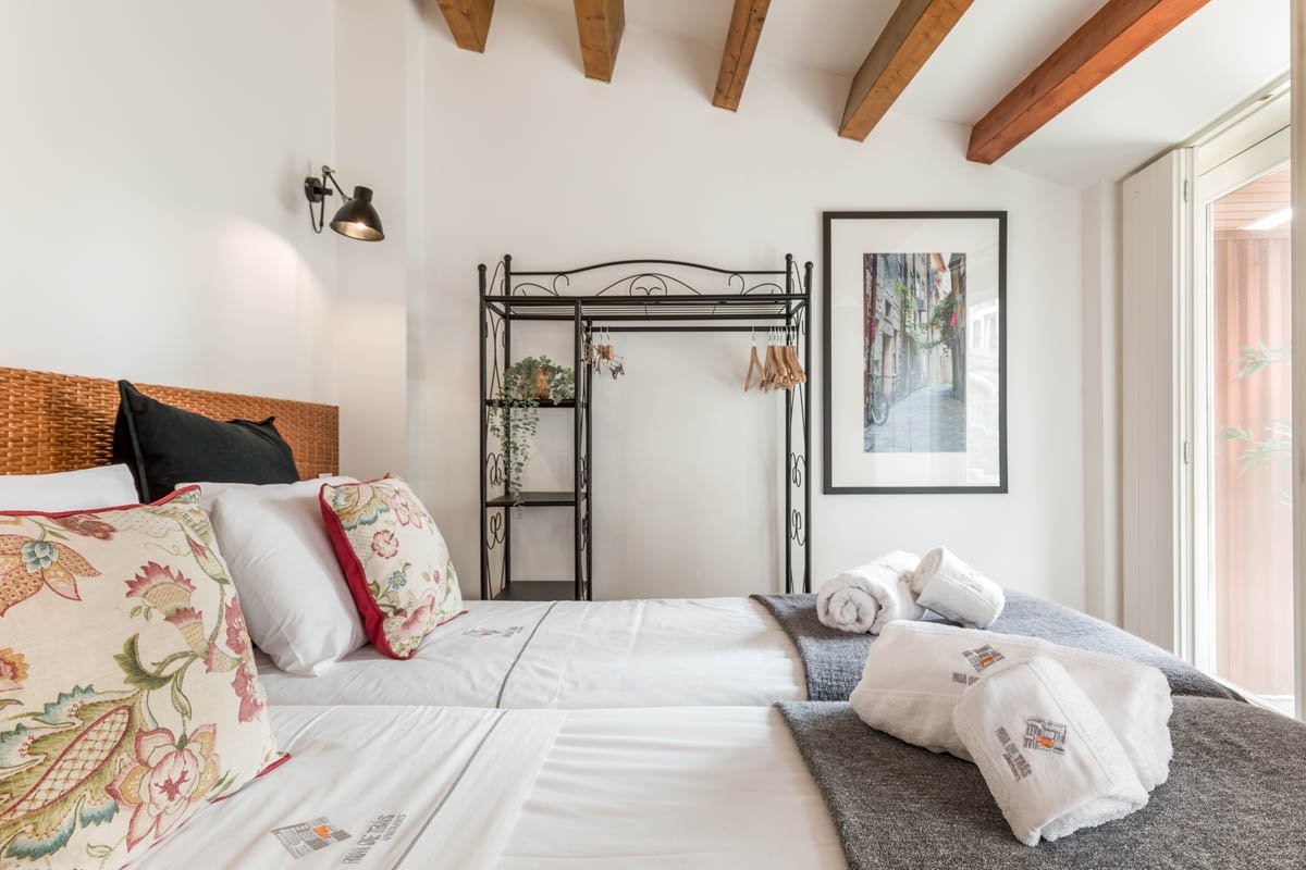 Quarto com duas camas de casal, cabeceira de palha, travesseiros decorativos florais, cobertores cinza, toalhas brancas dobradas, parede branca, lâmpada de parede, quadro com uma rua de paralelepípedos, janela grande com cortinas móveis, teto com vig