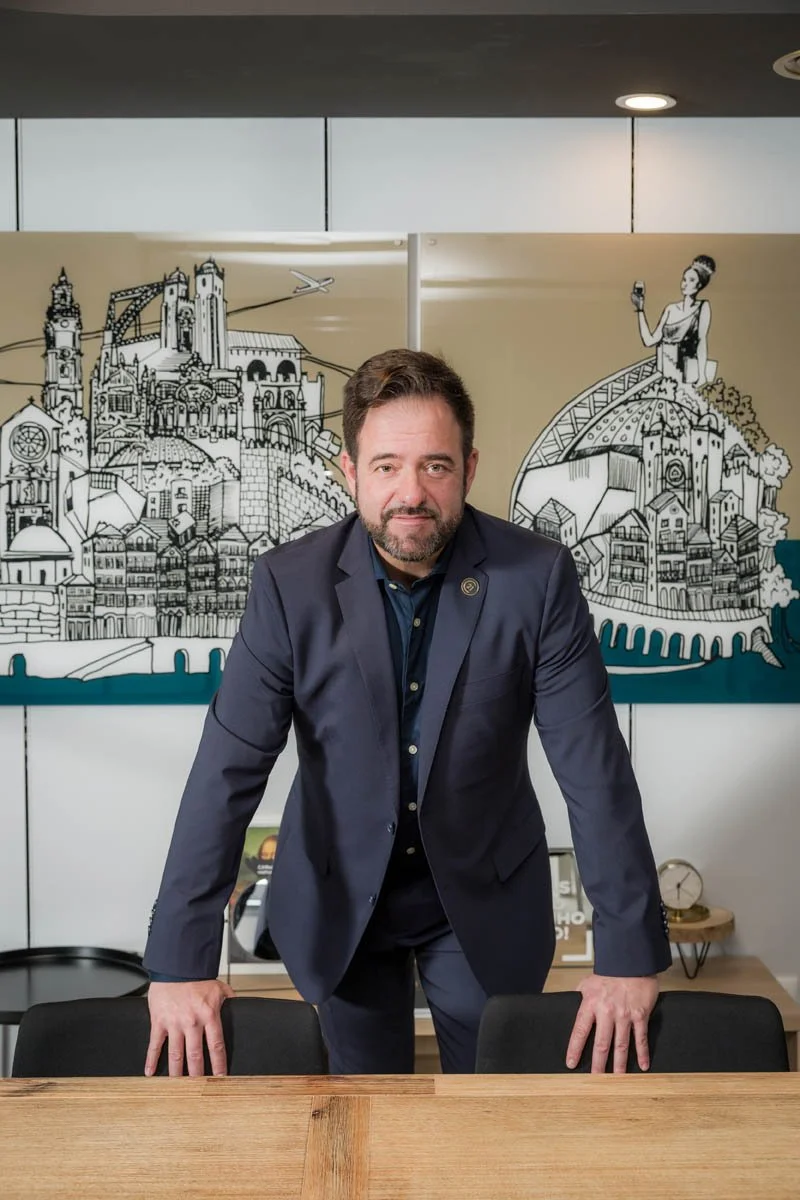 Homem de terno escuro, com barba, posando atrás de uma mesa de madeira em um escritório, com uma parede ao fundo que tem uma arte de desenho de uma cidade artística e uma mulher segurando uma taça.