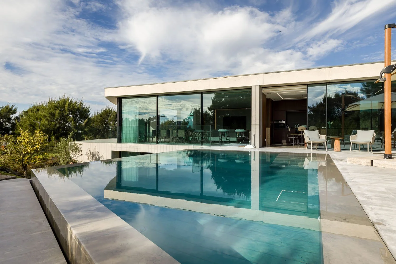 Imagem de uma casa moderna com piscina de água cristalina, varanda com cadeiras e jardim com árvores verdes, sob céu parcialmente nublado.