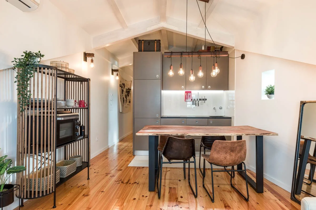 Cozinha moderna com teto inclinado, armários cinza, ilha de jantar com banquetas, prateleira de madeira com utensílios, prateleira com micro-ondas, iluminação com pendentes de filamentos, piso de madeira, parede branca e uma janela com planta.