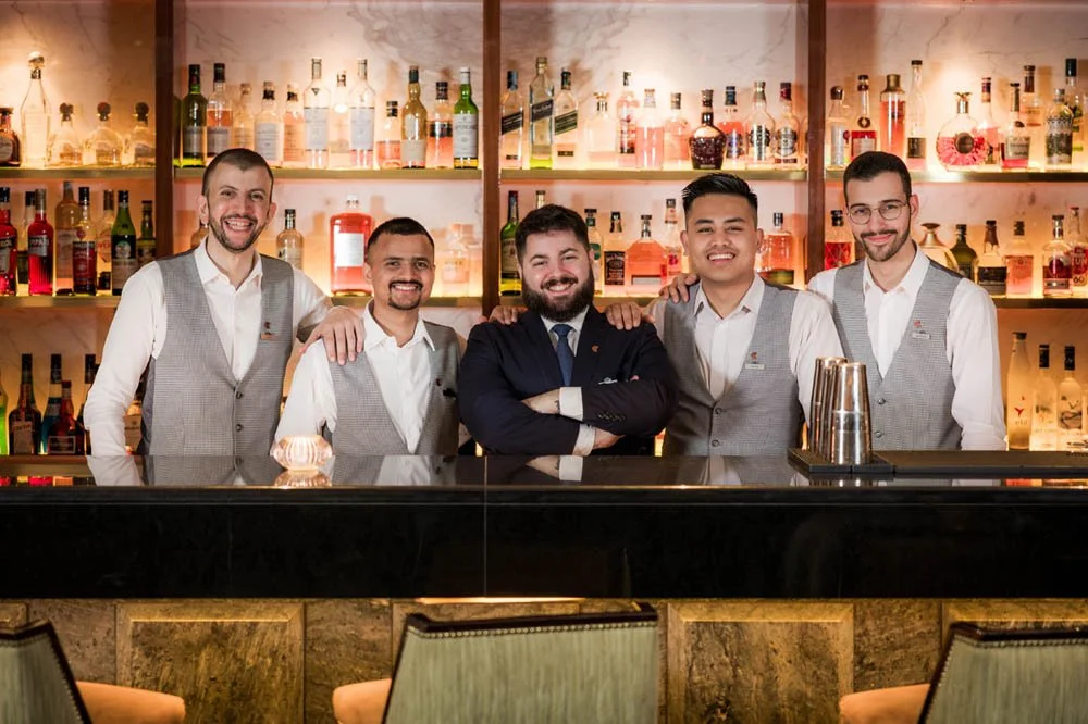 Seis barman sorridentes em um bar, com bebidas alcoólicas ao fundo.