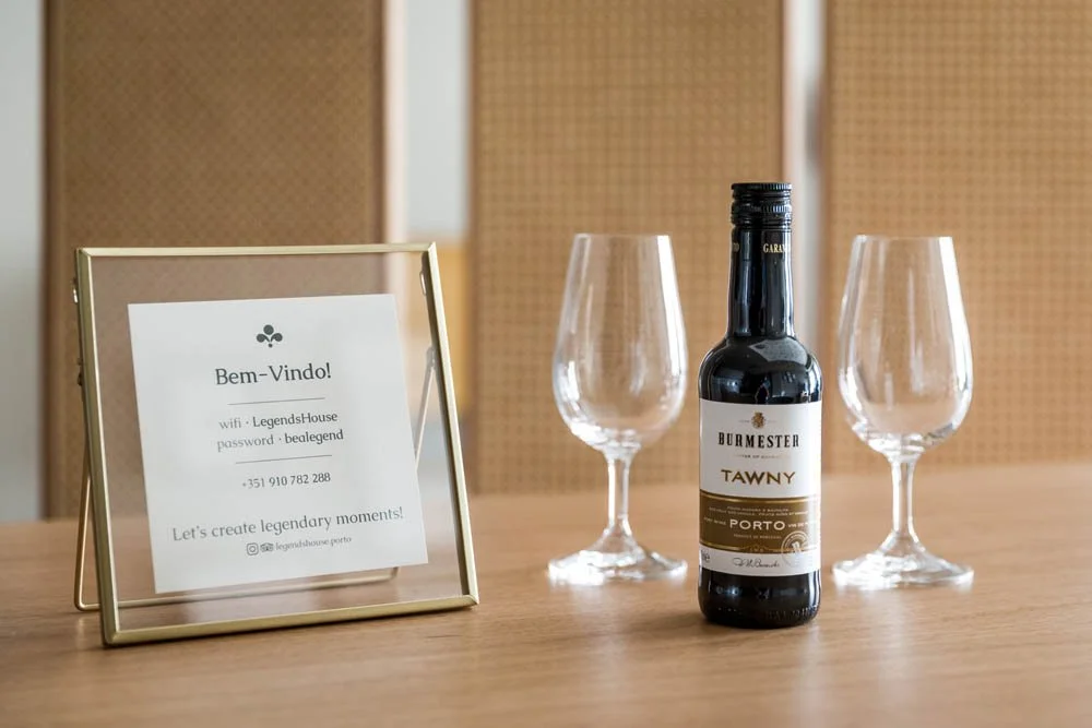 Uma garrafa de vinho tinto ao centro, ladeada por duas taças vazias de vinho, sobre uma mesa de madeira, com um quadro de boas-vindas ao lado com informações de login e telefone.