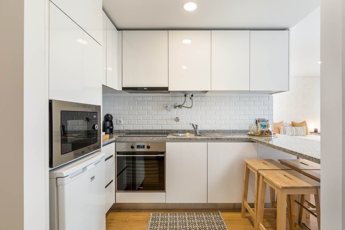 Cozinha moderna com armários brancos, pia de aço inox, fornos embutidos, micro-ondas, cafeteira, bancada de granito e duas banquetas de madeira ao lado.