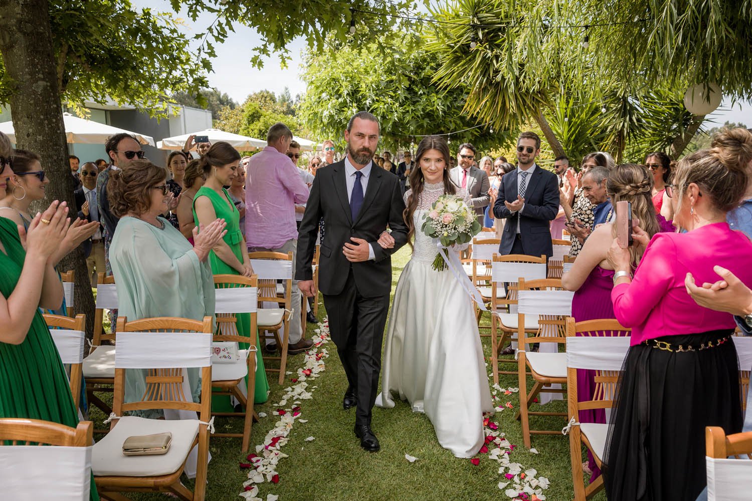 Casal de casamento caminhando pelo corredor ao ar livre, rodeado por convidados celebrando, com árvores e vegetação ao fundo.