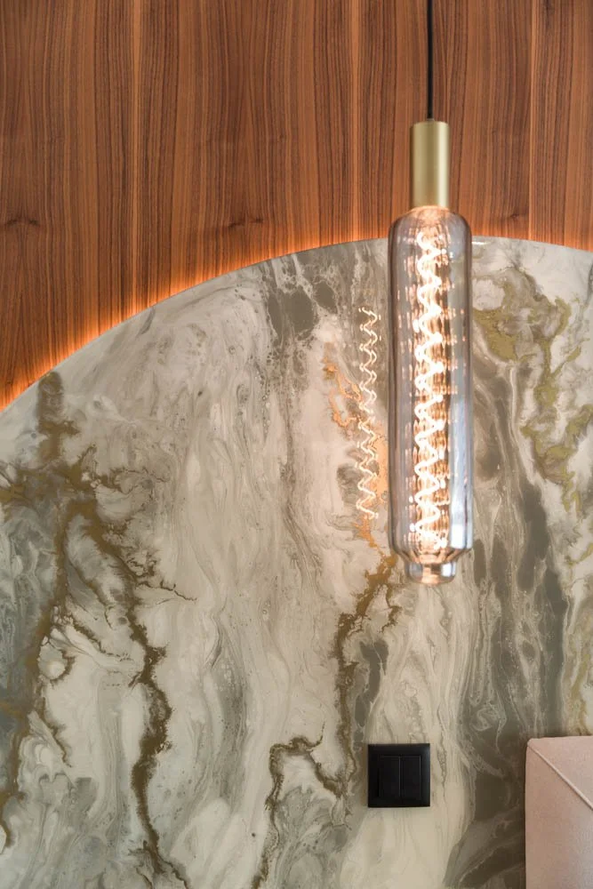 Luminária decorativa com lâmpada de filamento exposto, instalada em uma parede de mármore cinza com veios dourados, ao lado de uma parede de madeira com iluminação indireta.