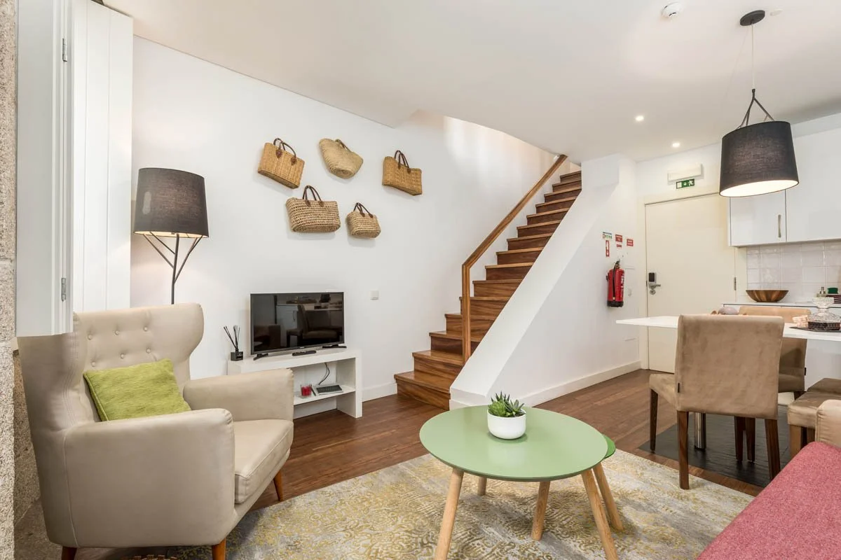 Sala de estar com sofá bege, cadeira com almofada verde, mesa de centro verde com planta, televisão na parede, luminária de chão ao lado do sofá, parede decorada com bolsas de palha, escada de madeira ao fundo, área de jantar com mesa branca e cadeir