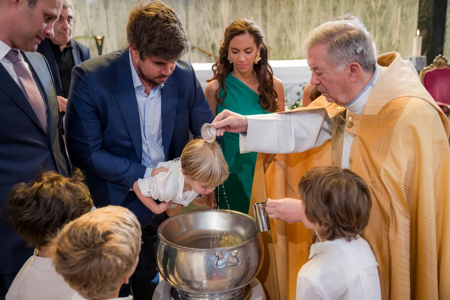 Papa católico abençoando uma criança com água benta durante uma cerimônia religiosa, rodeado por adultos e crianças.