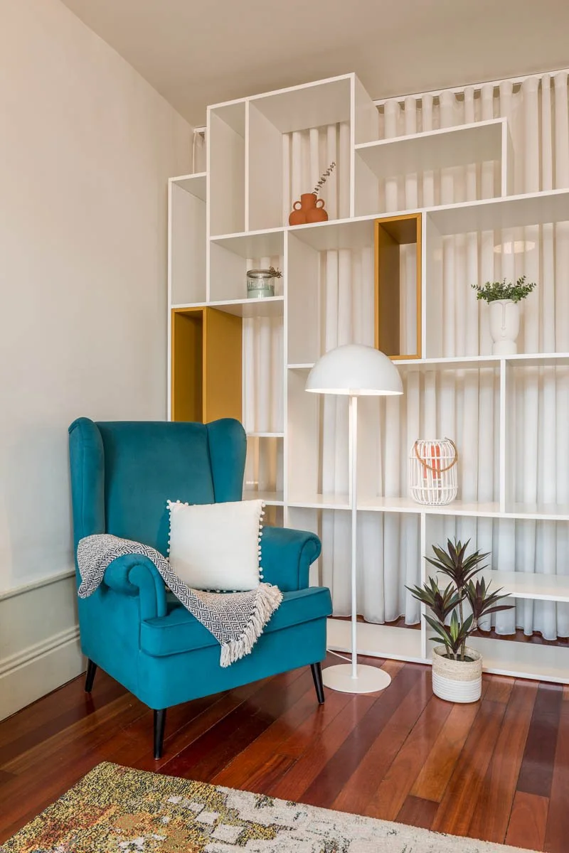 Poltrona azul com travesseiro branco e manta cinza, luminária de pé branca ao lado, prateleira branca decorada com vasos e objetos, piso de madeira, cortinas brancas ao fundo.