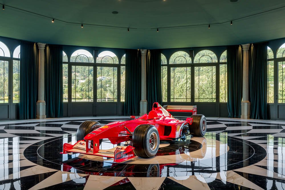 Carro de Fórmula 1 vermelho em uma sala de cristal com piso preto e branco e grandes janelas com cortinas.