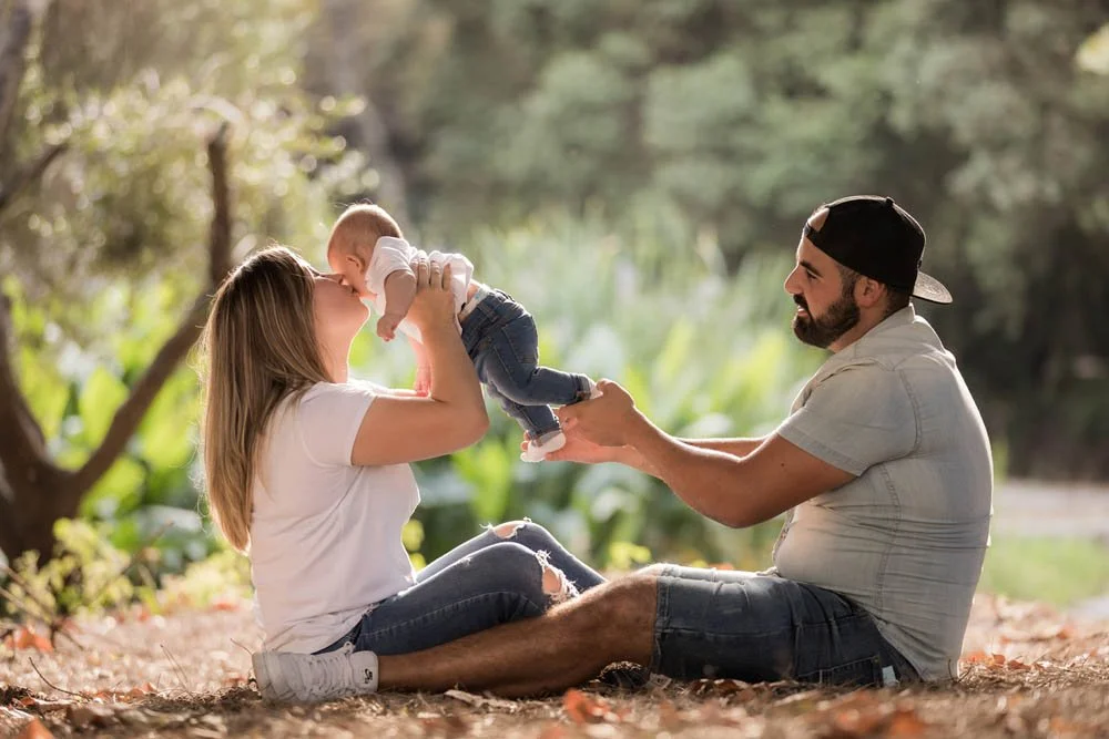 Família ao ar livre, mãe e pai brincando com o bebê sentado no chão, em um parque ou jardim, durante o dia com luz natural.