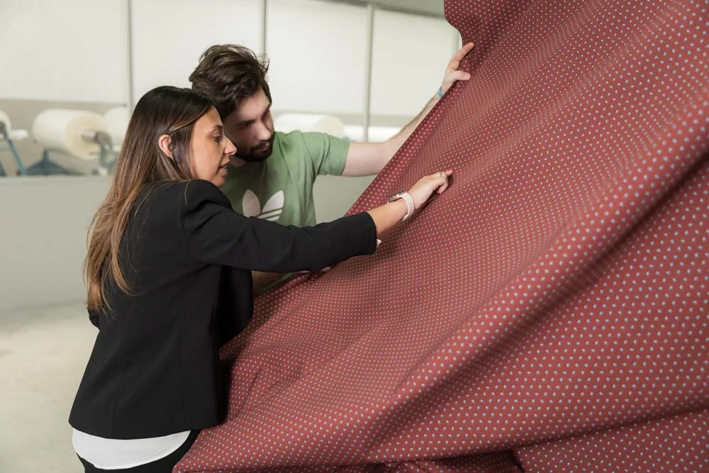 Homens e mulher examinam um tecido de cor vermelha com pontos brancos em uma fábrica ou loja de estofados.