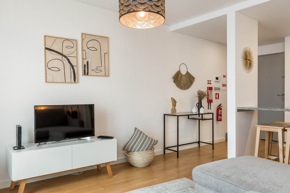 Sala de estar com televisão, quadros abstratos na parede, cômoda branca, decoração com espelho de palha, objeto de decoração de madeira, vaso com flores secas, relógio de sol dourado, parede branca e piso de madeira.