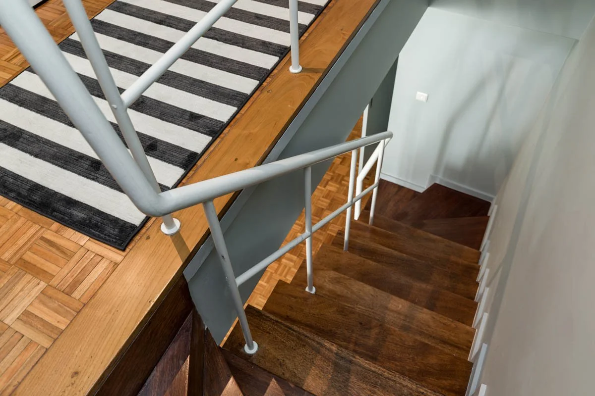 Escada de madeira com corrimão de ferro branco ao lado de uma sala com tapete listrado preto e branco.