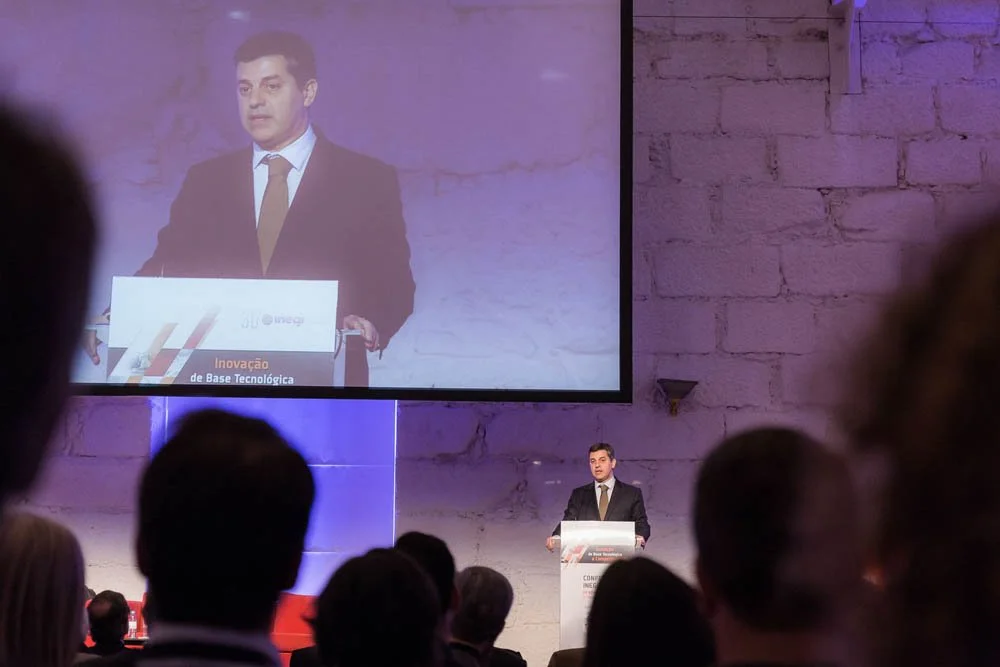 Homem de terno preto e gravata apresentando em um palco com público assistindo. Há uma tela grande acima, mostrando uma imagem ampliada dele em destaque na apresentação, com parede de tijolos ao fundo.