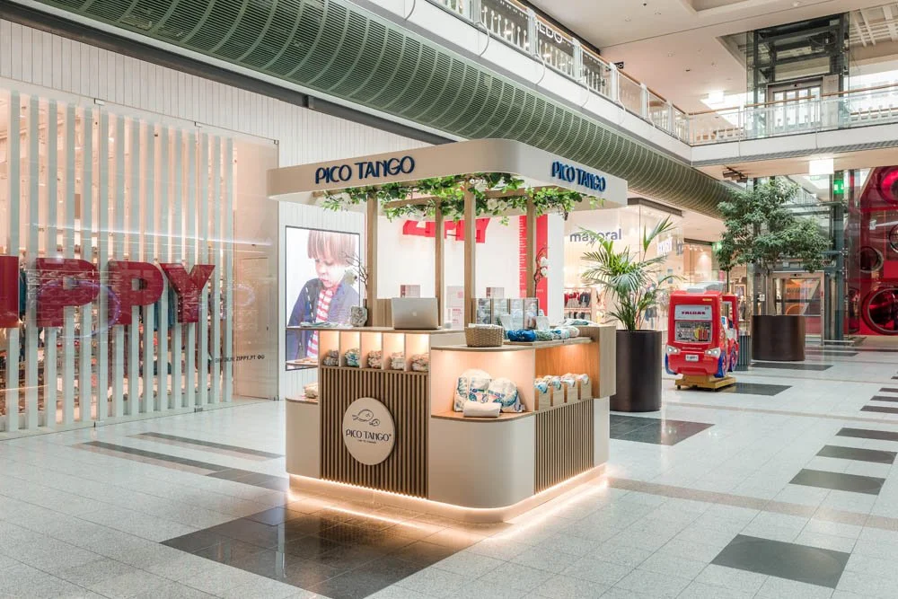 Quiosque de roupas infantis chamado Pico Tango em shopping, decorado com plantas, iluminação suave e brinquedo de madeira ao lado, ao fundo lojas e uma escada de acesso do shopping.