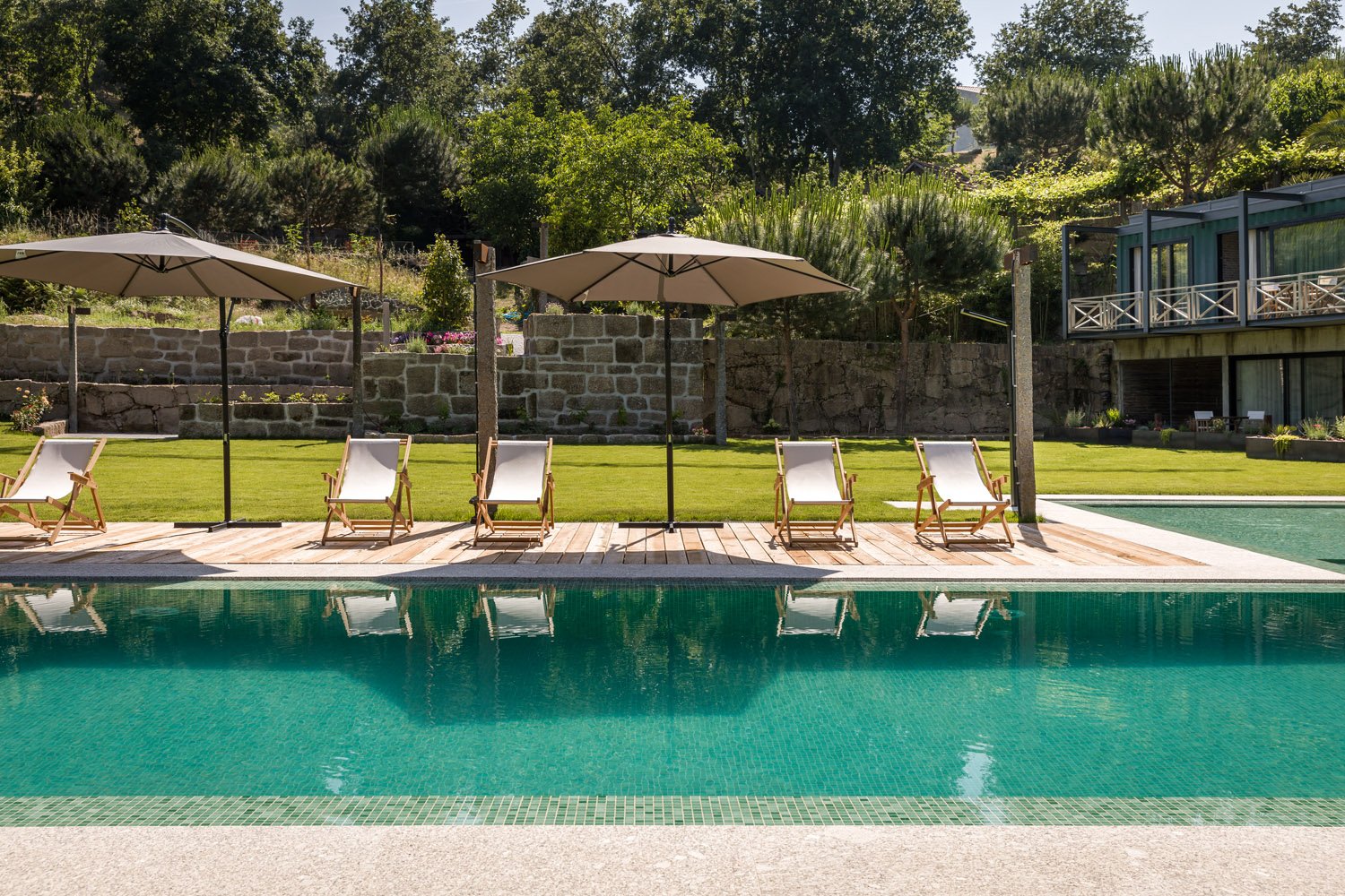 Piscina com espreguiçadeiras e guarda-sóis ao redor, jardim com grama verde e árvores ao fundo.