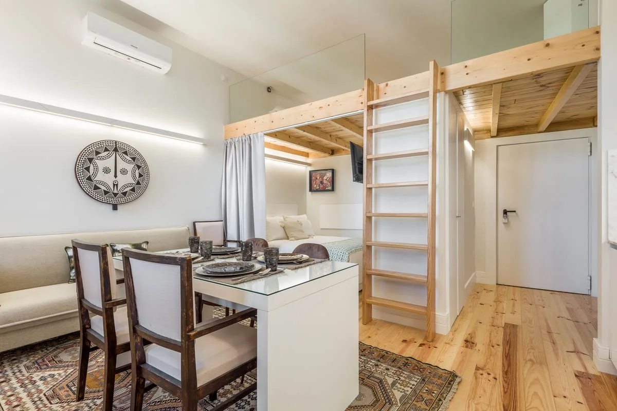 Sala de estar com área de dormir elevada separada por cortina, mesa de jantar com utensílios, parede decorada com peça de arte e televisão, piso de madeira e ar-condicionado.