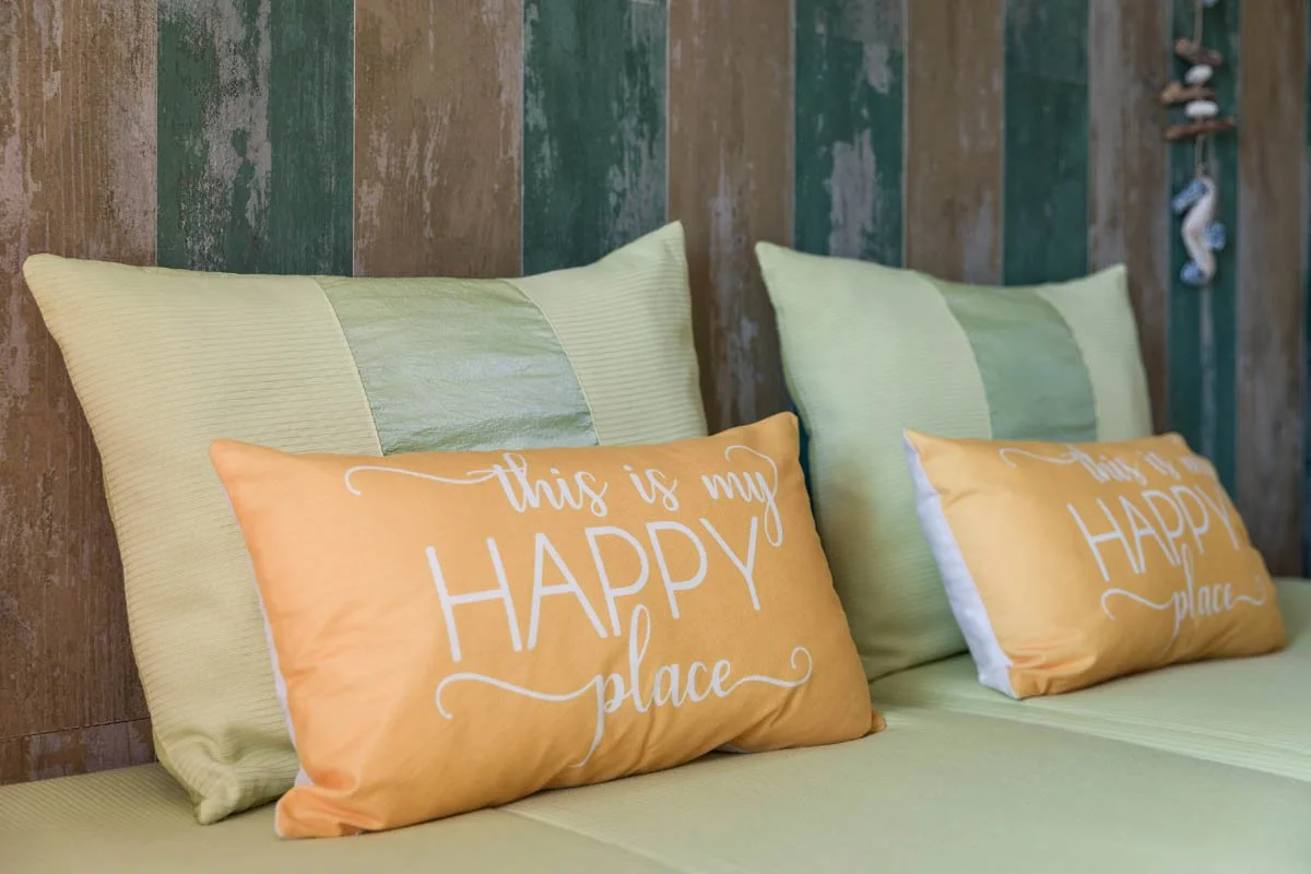 Cama com travesseiros verdes e almofadas laranjas com a frase "this is my happy place". Fundo de parede de madeira colorida.