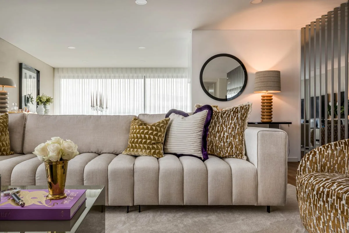 Sala de estar moderna com sofá bege, almofadas douradas e brancas, mesa de centro com vaso de flores brancas, espelho redondo na parede, luminária de mesa com base trançada e cortinas brancas ao fundo.