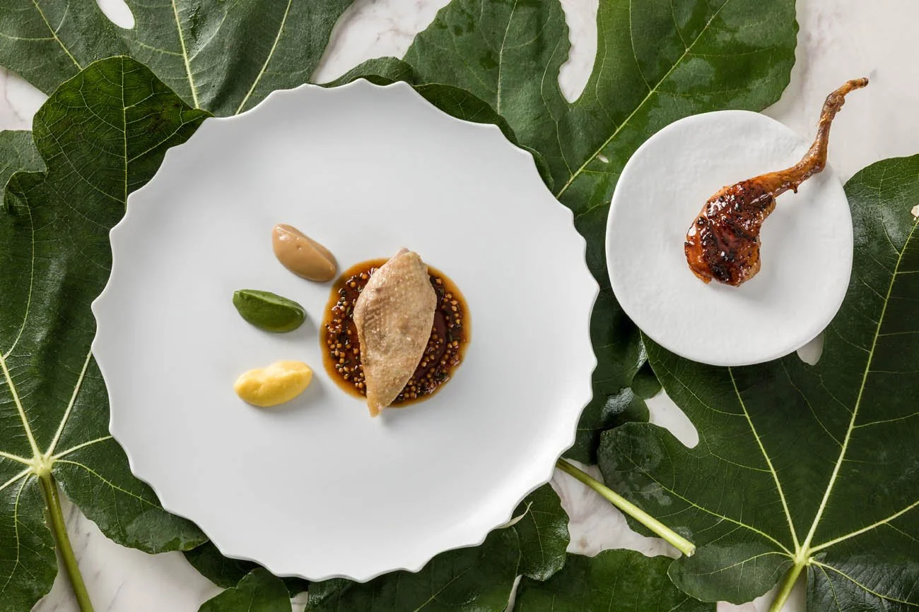 Prato elegante com fatia de carne ou peixe ao molho, acompanhada de três molhos ou guarnições, sobre louça branca com folhas verdes ao fundo, ao lado de ingrediente cozido ou grelhado em outro prato branco.