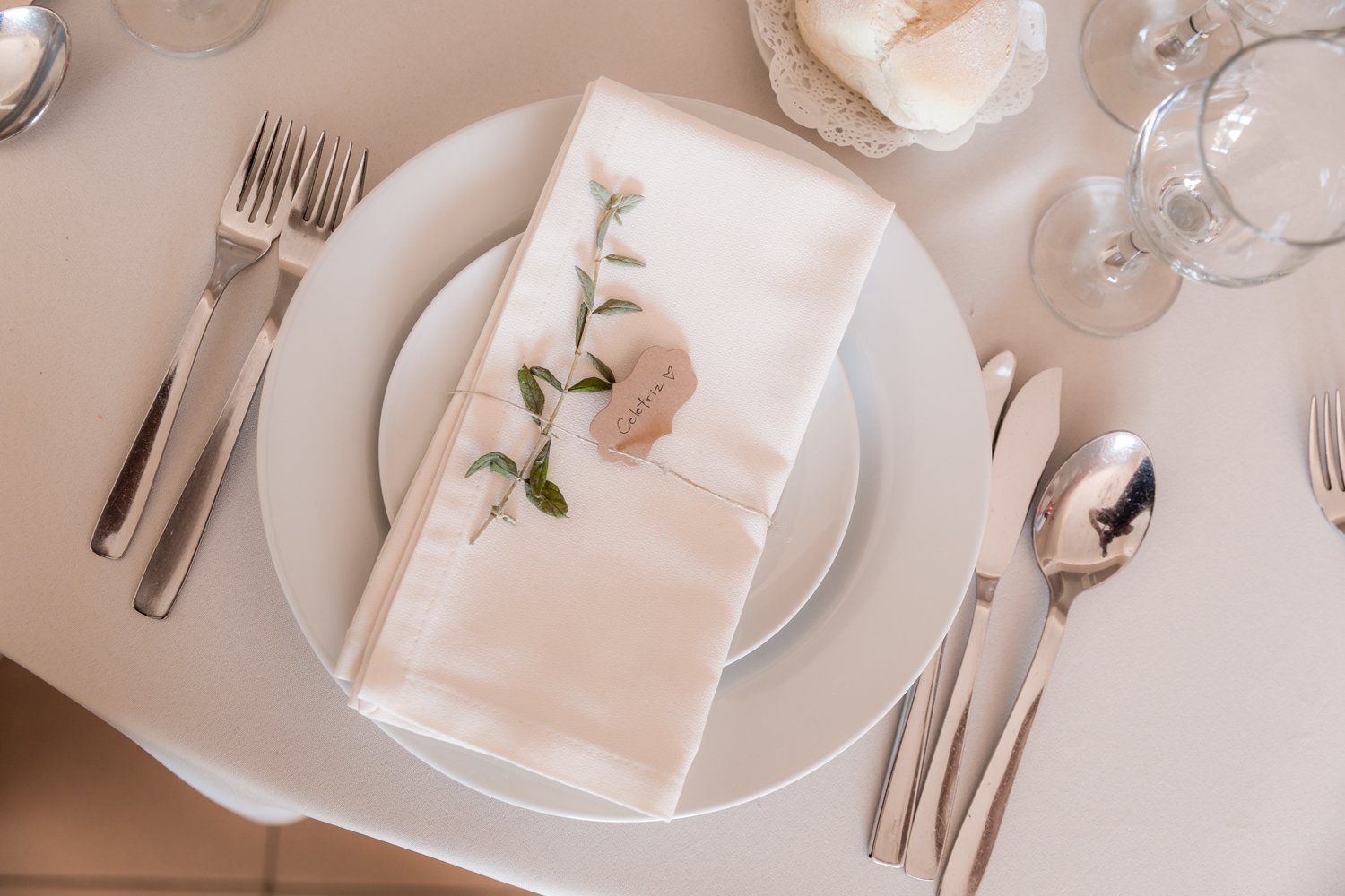 Configuração de mesa de jantar com prato, talheres de prata, guardanapo branco com decoração de galho de planta, uma pequena etiqueta com a palavra 'Coletiva', taças de vidro e pão em uma cesta