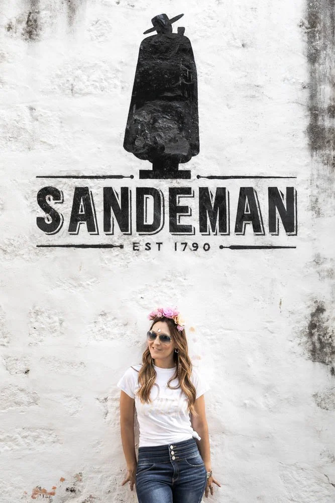 Mulher sorrindo usando óculos escuros, camiseta branca e jeans, com uma coroa de flores na cabeça, em frente a uma parede com logotipo da Sandeman e a frase "Est. 1790".