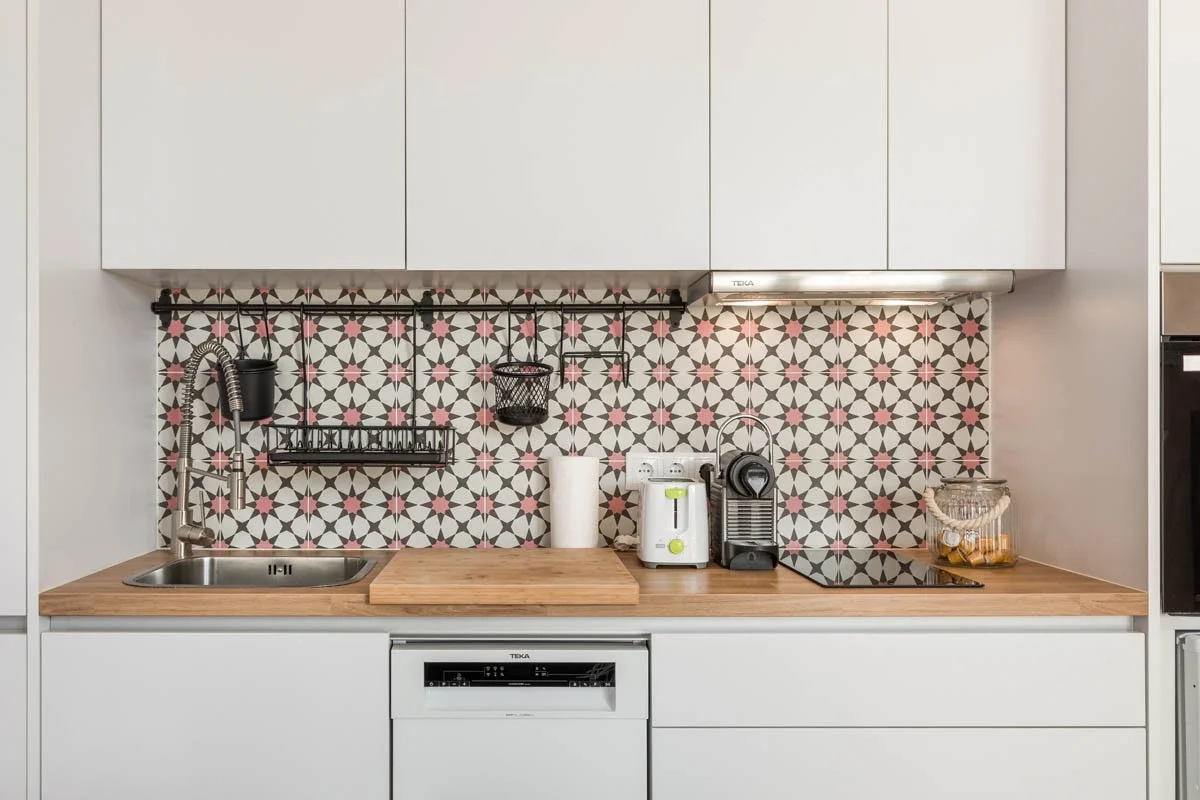 Cozinha moderna com backsplash decorativo em padrão geométrico rosa, preto e branco, com pia de aço inox, torneira de ponta alta, utensílios de cozinha, cafeteira, forno de micro-ondas embutido e fogão de vitrocerâmica.
