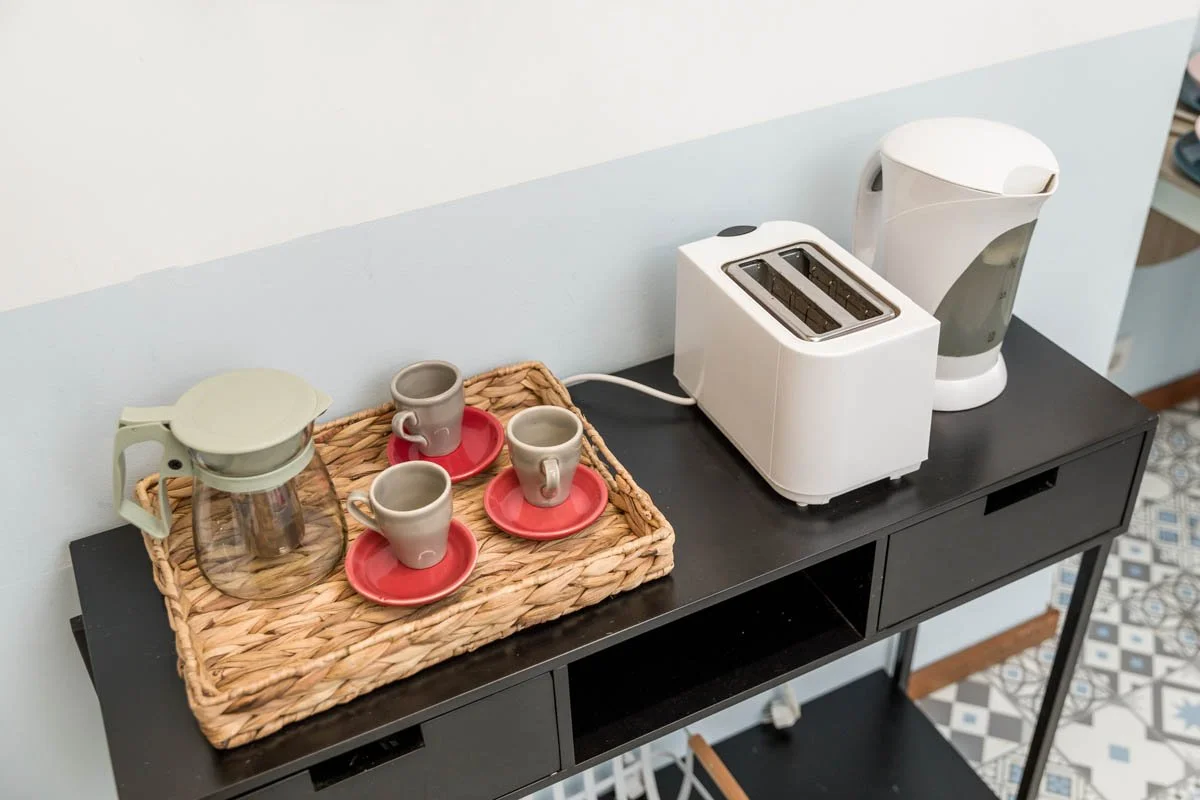 Balcão com uma cafeteira, um aparelho de água quente, uma torradeira branca, um recipiente com um jarro de vidro e três xícaras com suportes vermelhos, em ambiente de cozinha.