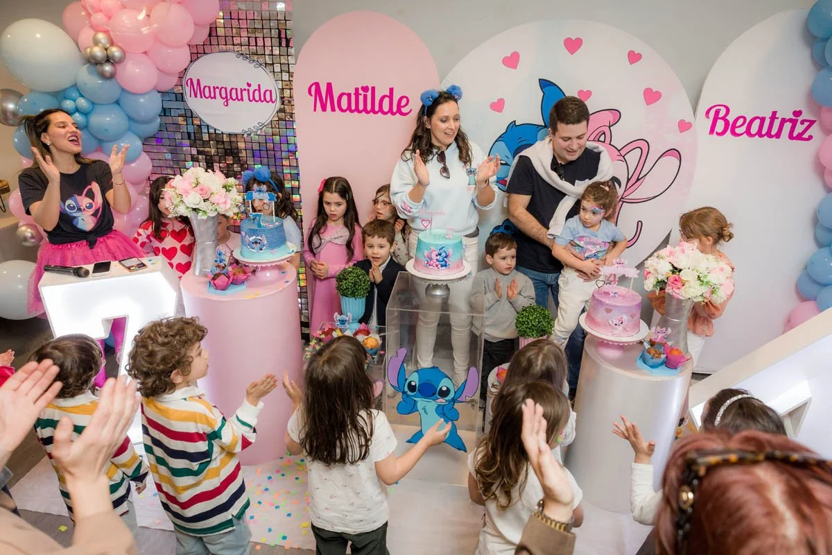Festa infantil com decoração rosa, azul e branca, com balões, flores e personagens do desenho 'Stitch'. Crianças estão celebrando, com mesas de doces, bolos decorados e convidados comemorando.