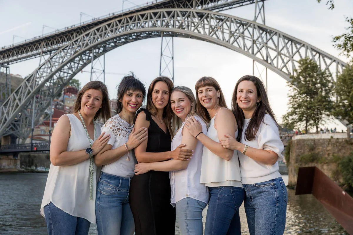Grupo de seis mulheres sorrindo na frente de uma ponte metálica ao lado de um rio.