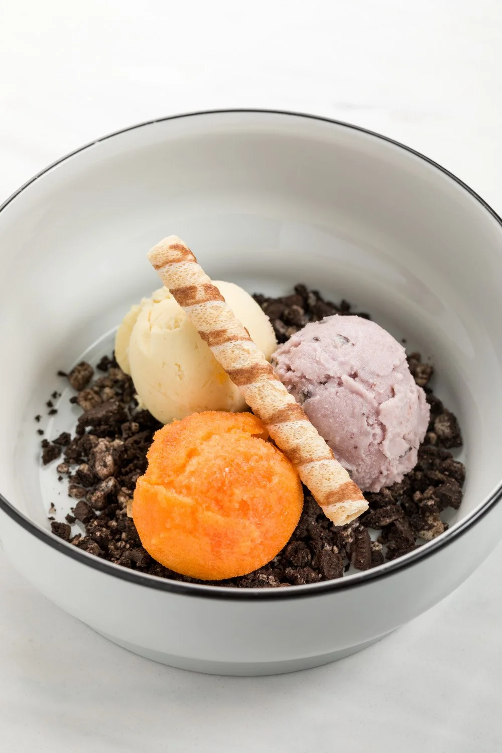 Taça com três bolas de sorvete de cores diferentes, pedaços de biscoito, pedaços de chocolate e um biscoito no topo.