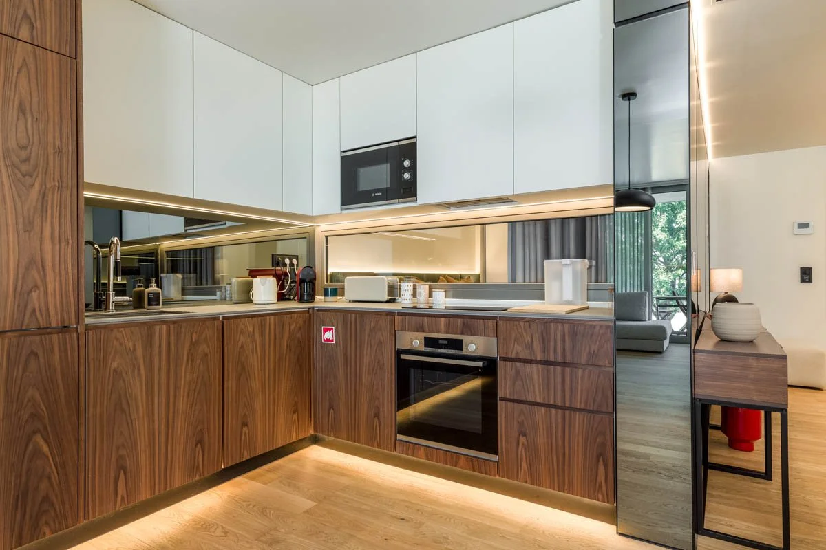 Cozinha moderna com mobiliário de madeira e armários brancos, eletrodomésticos embutidos, parede de vidro que reflete a sala de estar com sofá e luminária.