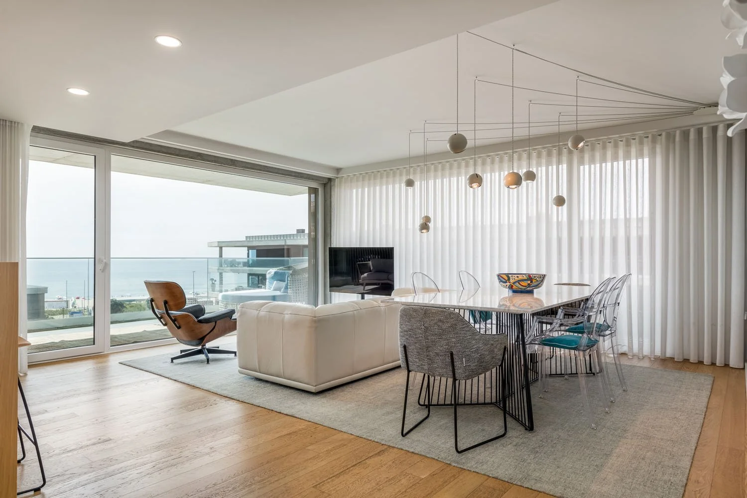 Sala de jantar moderna com paredes de vidro e vista para o mar, móveis de design contemporâneo, incluindo uma mesa de mármore com cadeiras transparentes, um sofá branco, uma poltrona de estilo escandinavo e iluminação pendente com globos