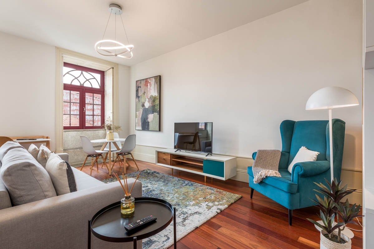 Sala de estar com sofá cinza, poltrona azul, tapete multicolorido, mesa de centro preta, televisão em móvel branco e marrom, janela grande com moldura vermelha, luminária moderna no teto, quadro decorativo na parede, planta em vaso e luminária de chã