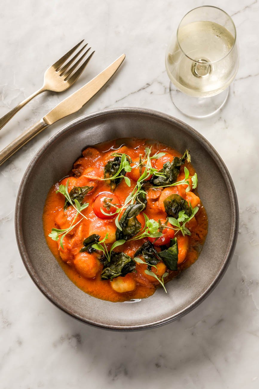 Prato com molho de tomate e folhas verdes, ao lado de uma taça de vinho branco e talheres dourados em uma superfície de mármore.