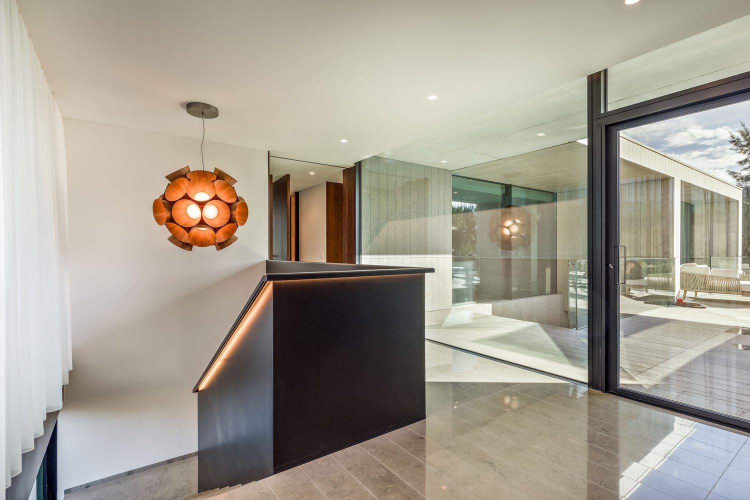 Interior de uma casa moderna com escada preta, iluminação embutida e uma grande janela de vidro, com área externa visível com móveis.