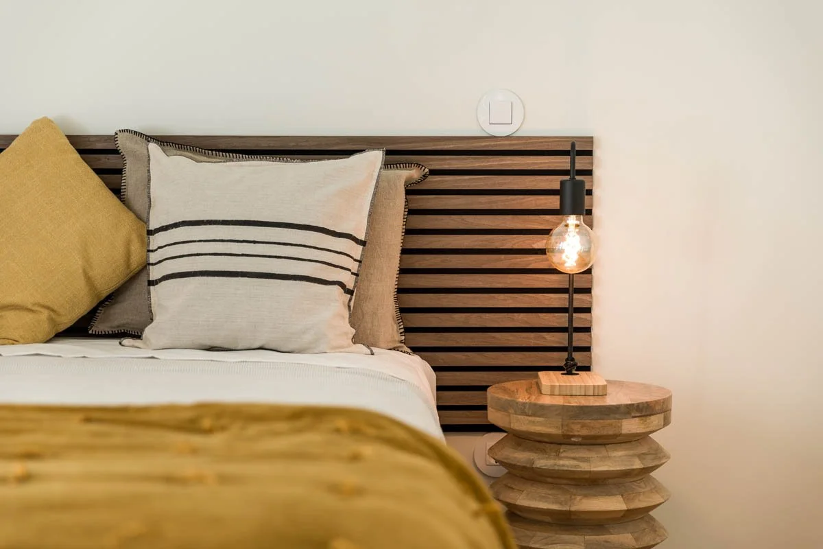 Cama com almofadas de cores amarela, branca com listras pretas, e bege, ao lado de um criado-mudo de madeira com um abajur de lâmpada exposta